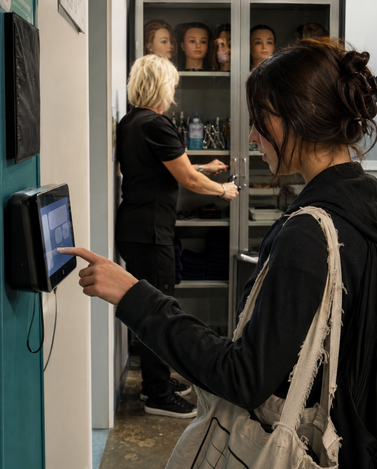 Cosmetology student records her arrival on an attendance terminal while an instructor prepares mannequins and sanitized tools for class.