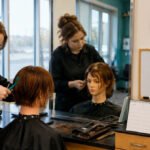 Cosmetology student practices a layered mannequin haircut beside a clock, calendar, and blank board used to track required training hours.