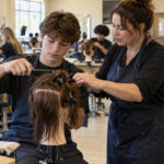 High school students practice comb control on training mannequins while an adult teacher demonstrates cosmetology techniques in a vocational classroom.