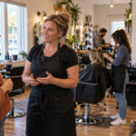 Prospective cosmetologist speaking with an experienced stylist while supervised salon training and mannequin-based classroom practice continue nearby.